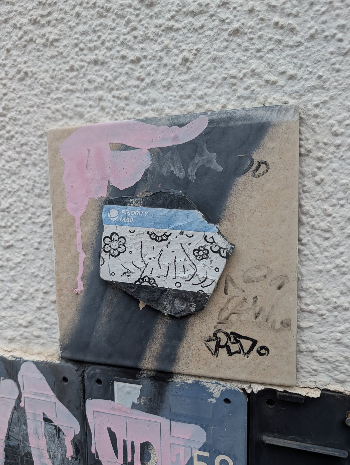 Quadratische helle Fliese mit schwarzer Verlaufsfläche und rosa Farbspritzern auf einer strukturierten weißen Wand. Mittig klebt Priority-Mail-Aufkleber, der mit schwarzen Blumen- und Linienzeichnungen bemalt ist.