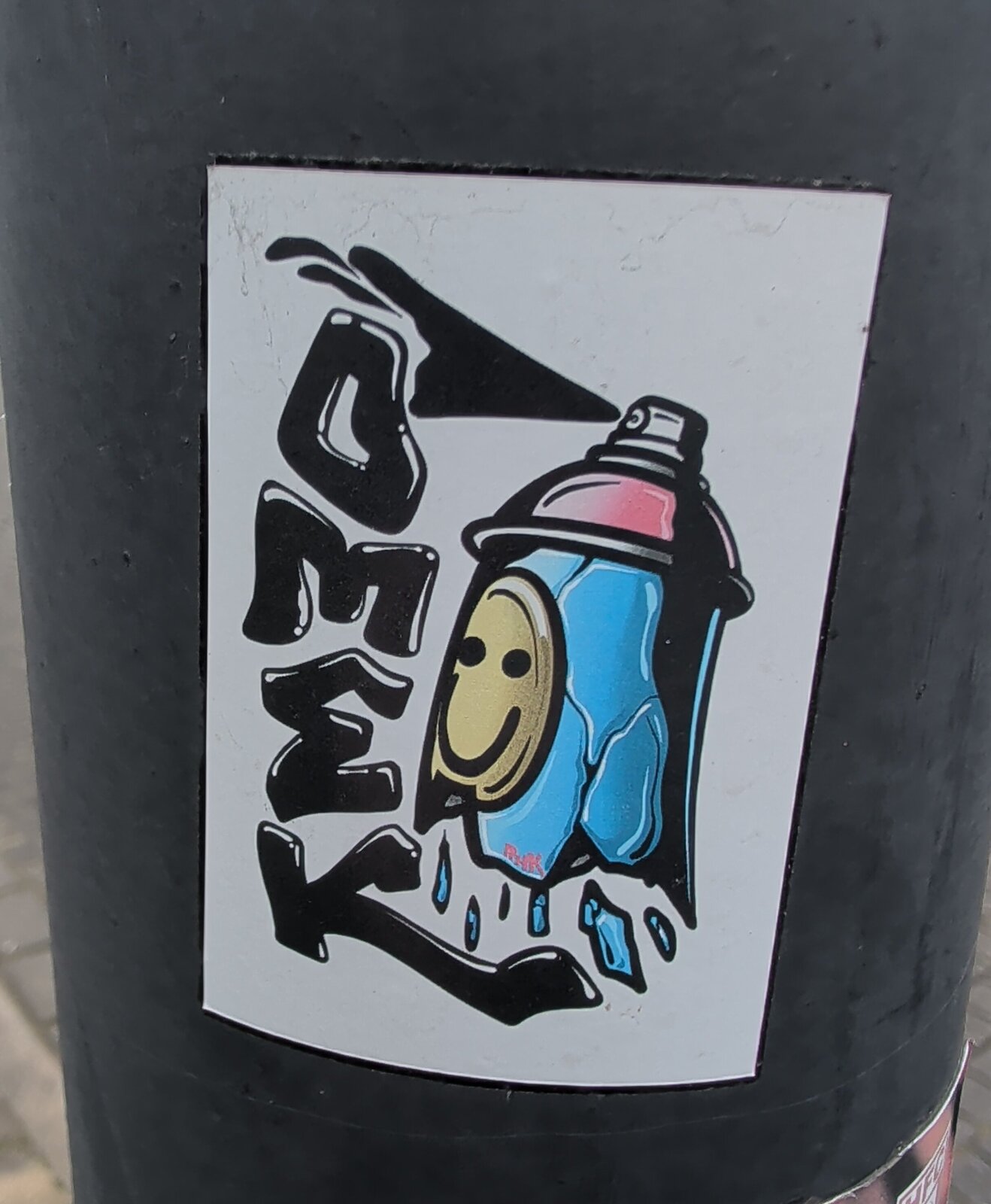 Aufkleber auf einem dunklen Metallmast: schwarze, stilisierte Schrift mit einem Pfeil, rechts eine blau-rosa Spraydose mit gelbem Smiley-Gesicht, aus der Farbe tropft. Weißer Hintergrund. Geschrieben steht OMEK.