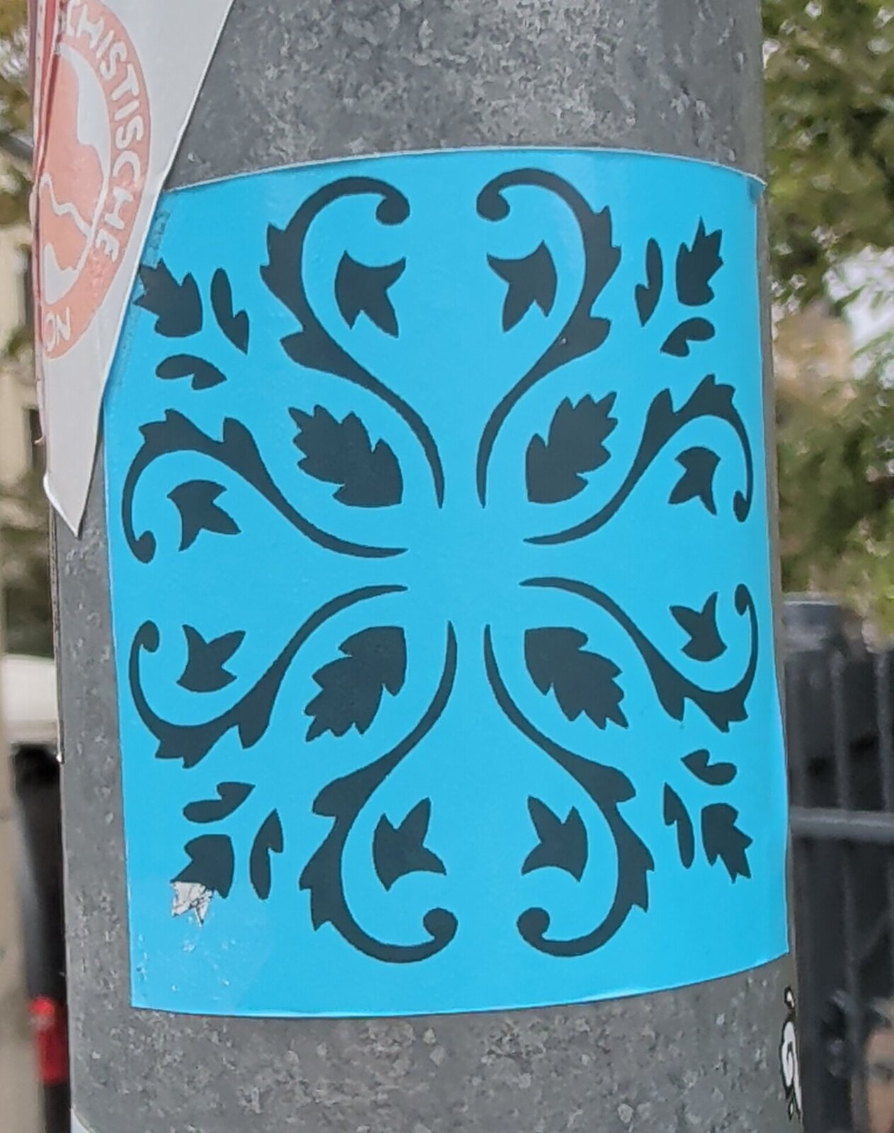 Blauer Aufkleber mit dunkelblauem, symmetrischem Ornament aus Ranken und Blättern auf einem grauen Laternenmast.
